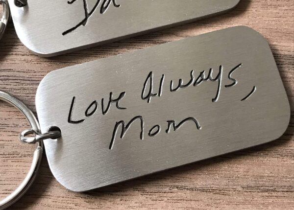 Engraved keychain with handwritten message Love Always, Mom on a metal tag, resting on a wooden surface.