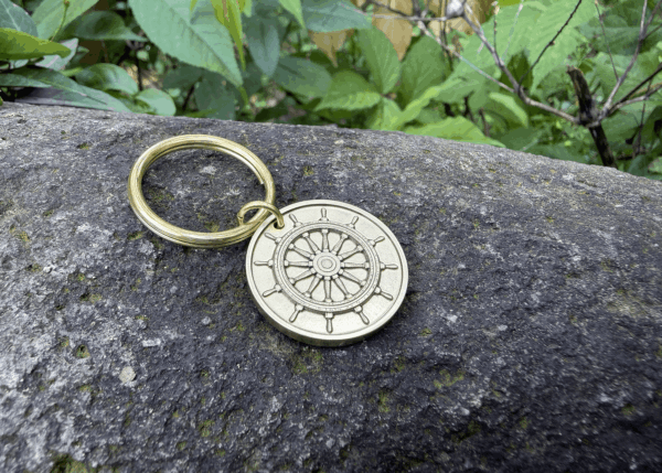 Gold keychain with nautical ship wheel design on textured rock, surrounded by green foliage.