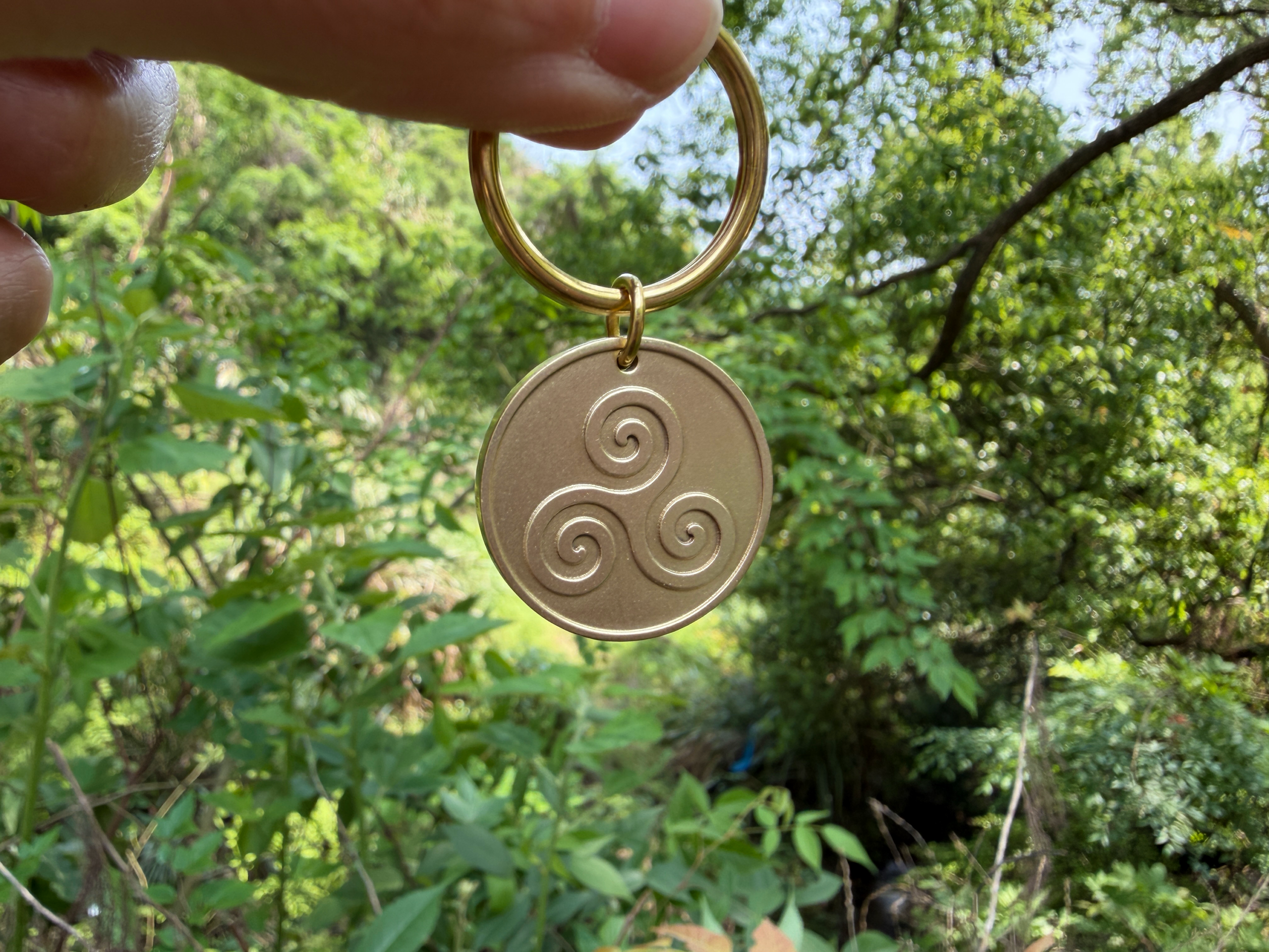 A person holds a keychain with a triskelion engraving, set against a lush green forest background.