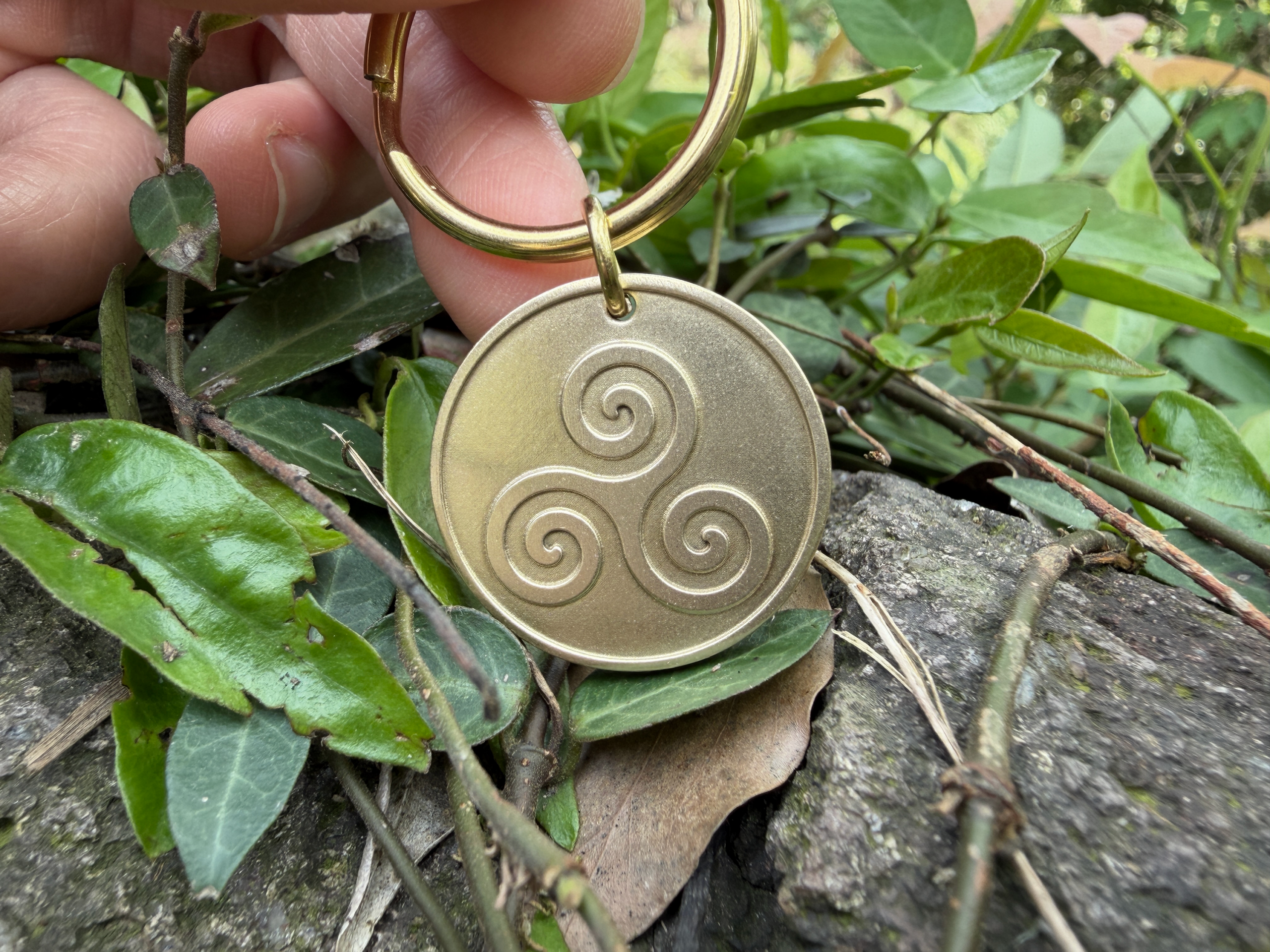 Gold keychain with Celtic triple spiral engraving, known as a triskele, held over green leaves and stone background.