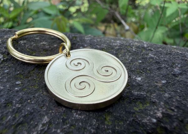 Gold keychain with a triskelion engraving on a stone surface; green foliage background.