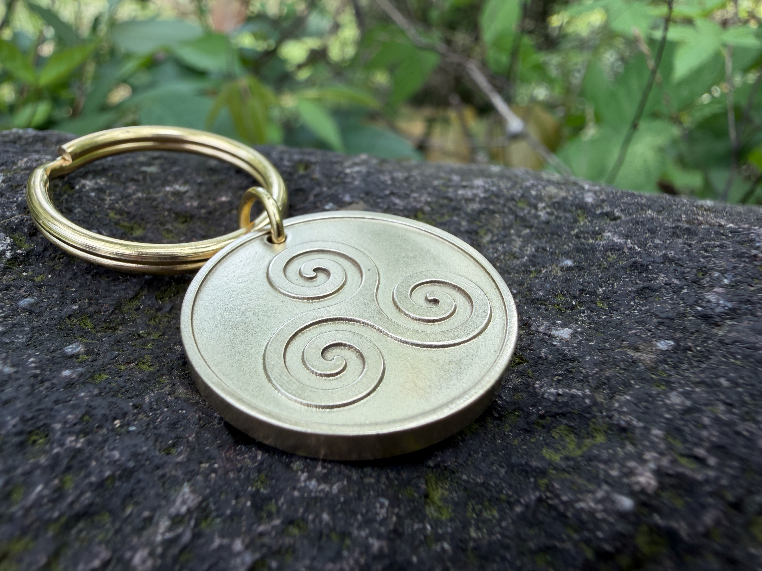 Gold keychain with a triskelion engraving on a stone surface; green foliage background.