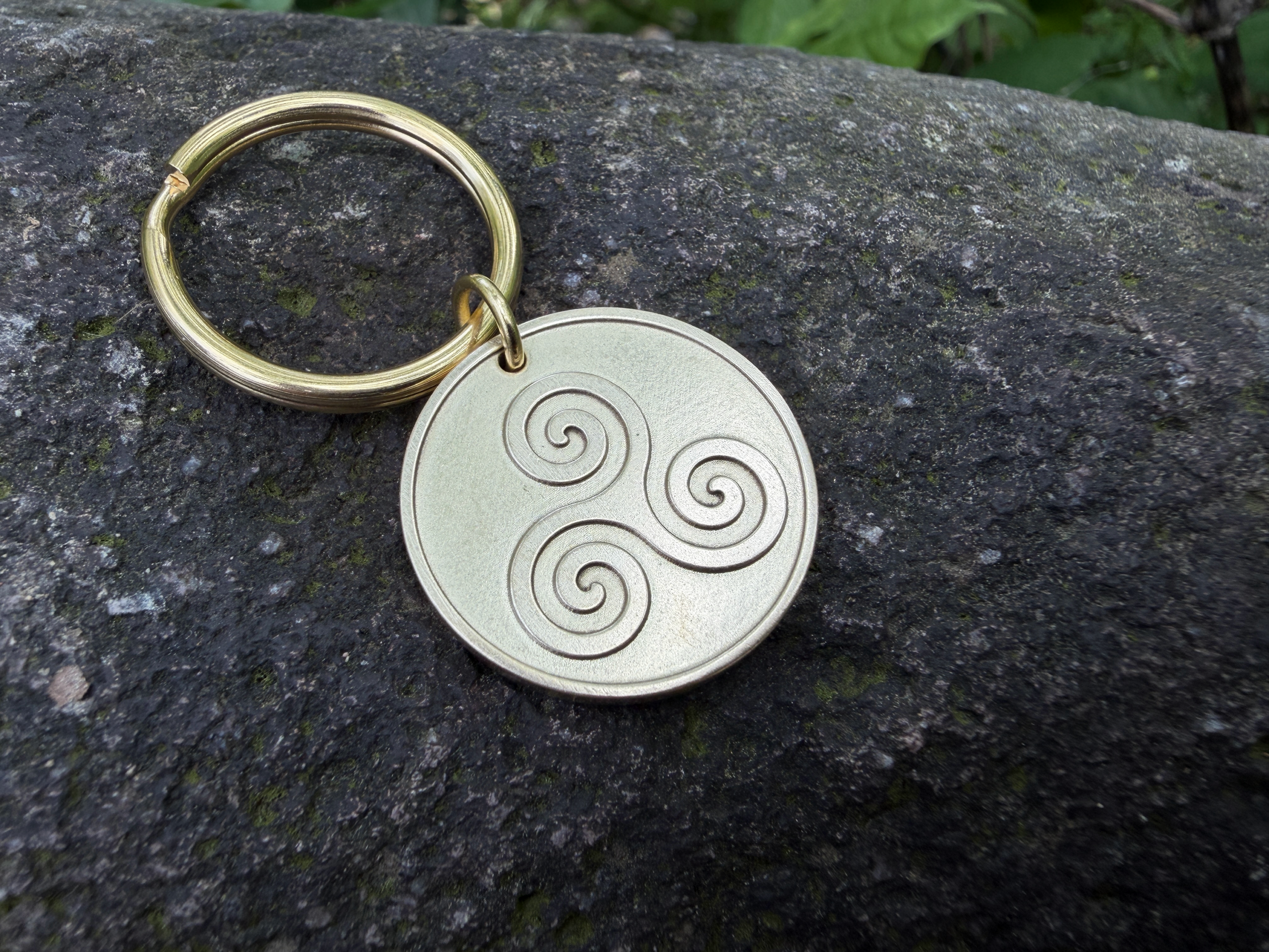 Gold keychain with Triskelion engraving on a stone background, symbolizing motion and progress. Celtic-inspired design.