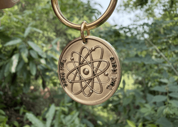 Gold keychain with atomic symbol engraving and customizable text in a lush green outdoor setting.
