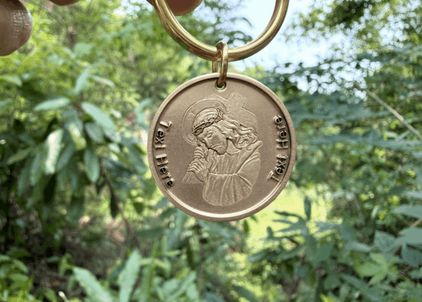 Gold medallion keychain with Jesus Christ engraving, held against a lush green background. Text unfinished.