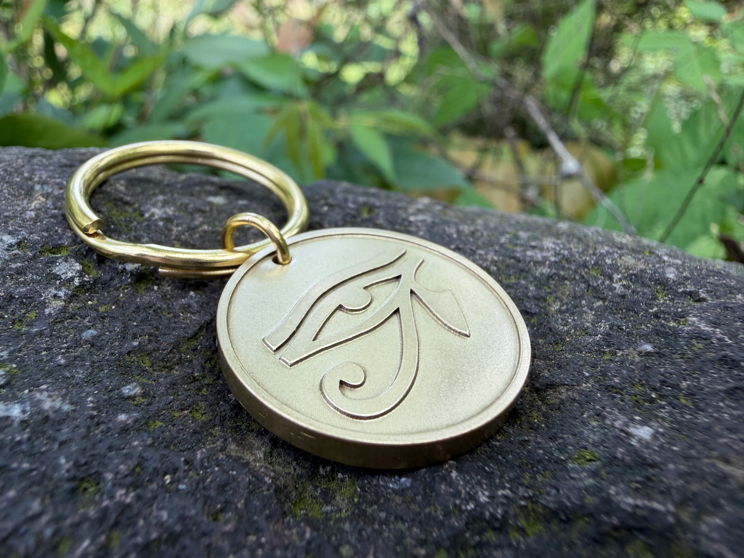 Golden keychain with Eye of Horus engraving on rock surface, surrounded by greenery in the background.