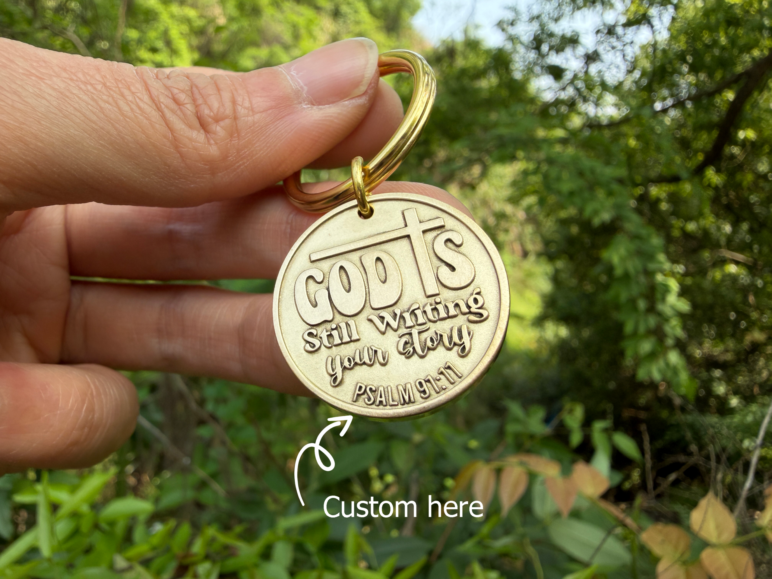 Keychain with God is still writing your story engraved, held in hand, nature background, Psalm 91:11 reference.