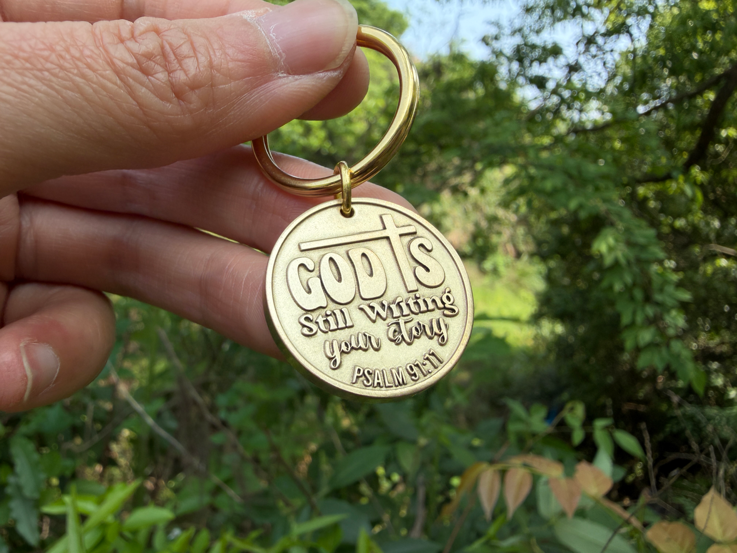 Gold keychain with engraving God's Still Writing Your Story, Psalm 91:11, held in nature background.