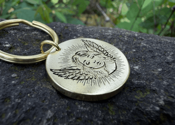 Gold keychain with an engraving of a baby angel on a textured stone surface.