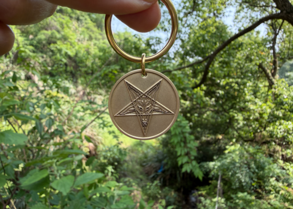 Pentagram keychain with goat engraving, held in nature setting, surrounded by lush green trees.