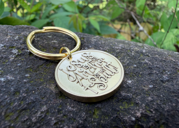 Gold keychain on stone with engraving Jesus is the Anchor of my Soul, set against lush green foliage background.