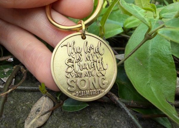 Keychain engraved with The Lord is my strength and my song, Psalm 117:13, held outdoors near leaves.