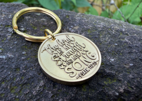 Gold keychain engraved with The Lord is my strength and my song - Psalm 118:14 on a stone background.