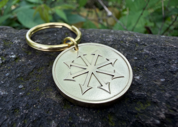 Gold metal keychain with chaos star engraving resting on a rock with greenery in the background.