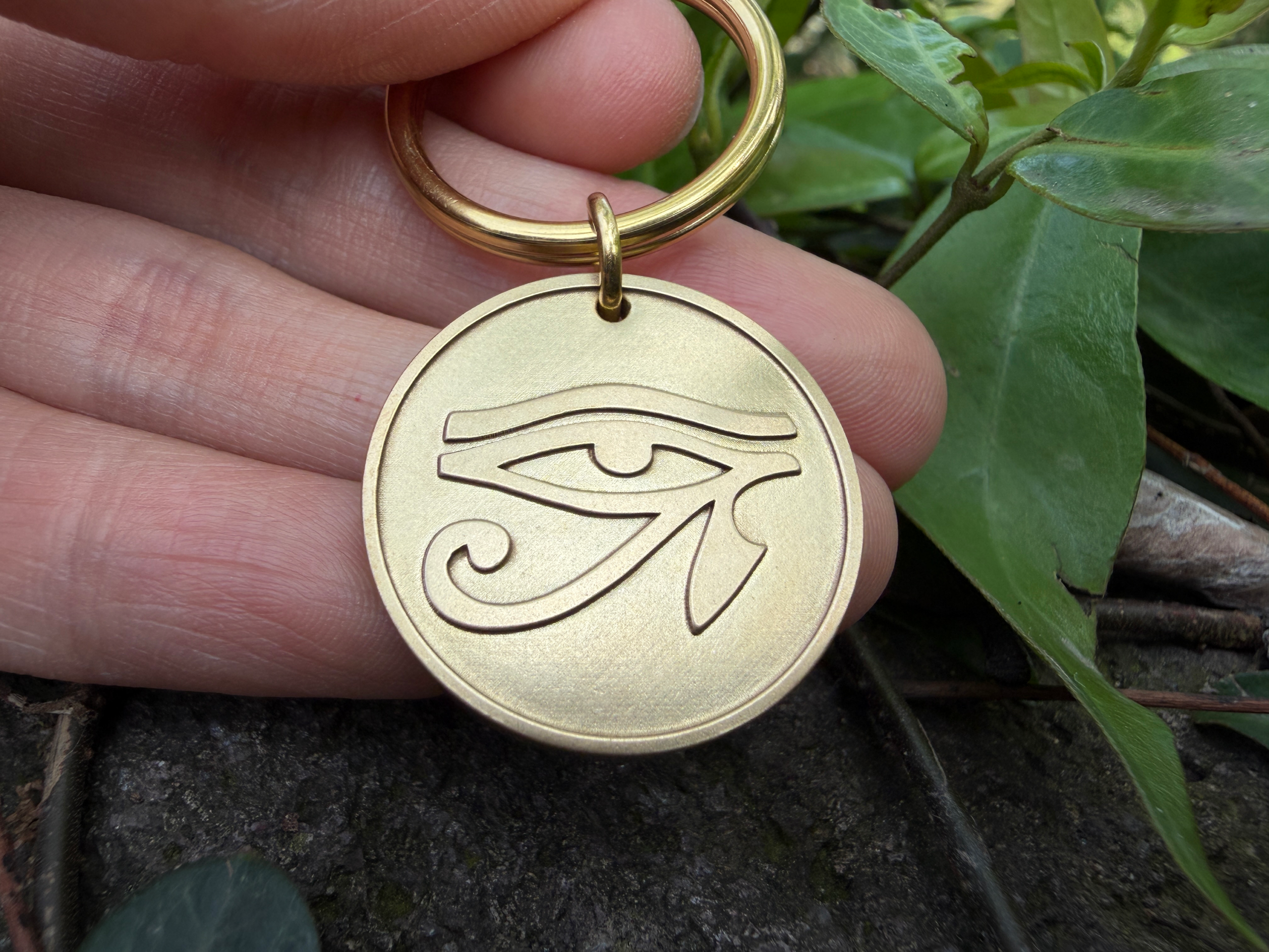 Golden keychain engraved with Eye of Horus held between fingers amid green foliage.