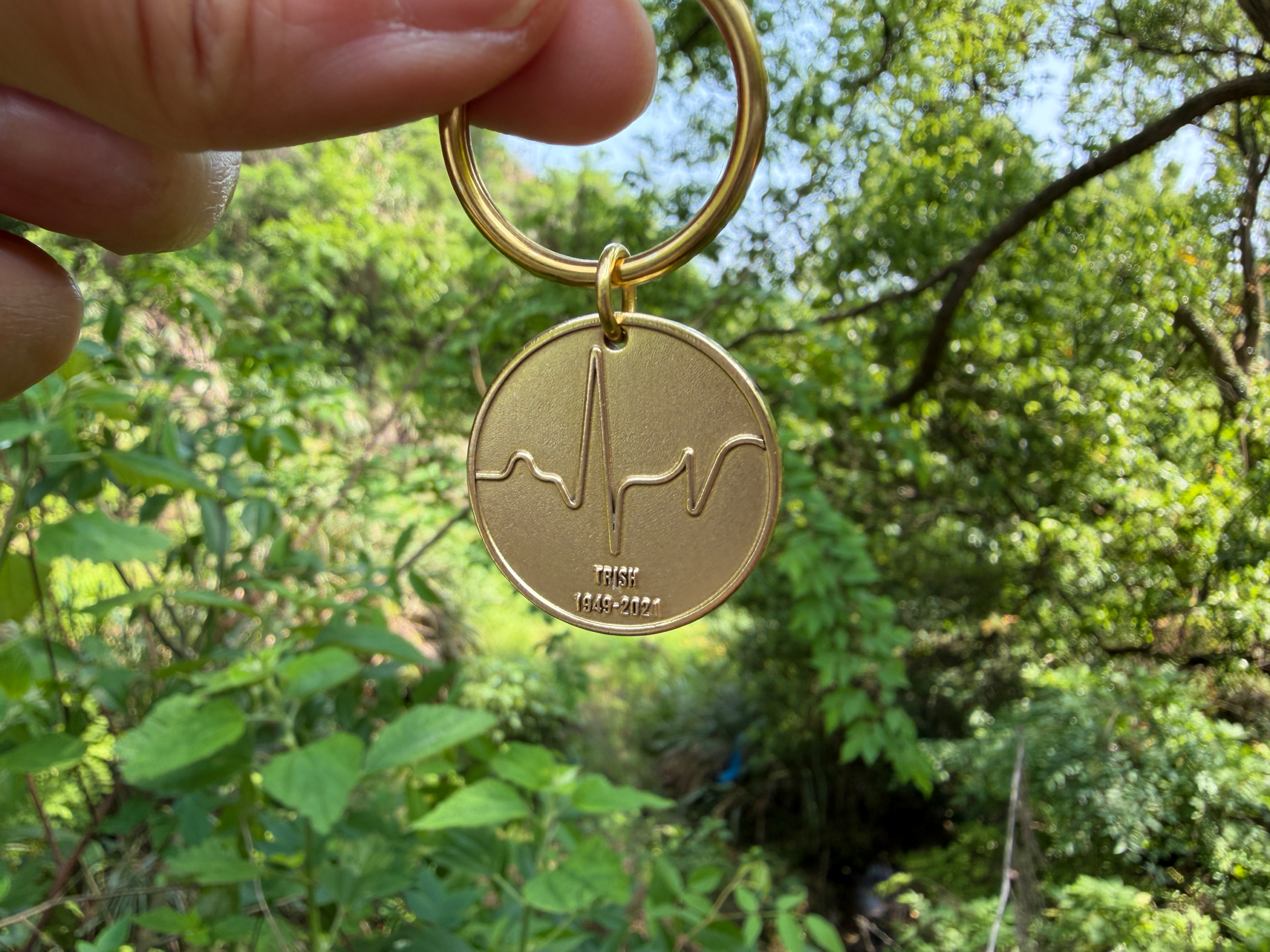 Gold keychain with heartbeat engraving, memorial text TRISH 1949-2021, held against a green leafy background.