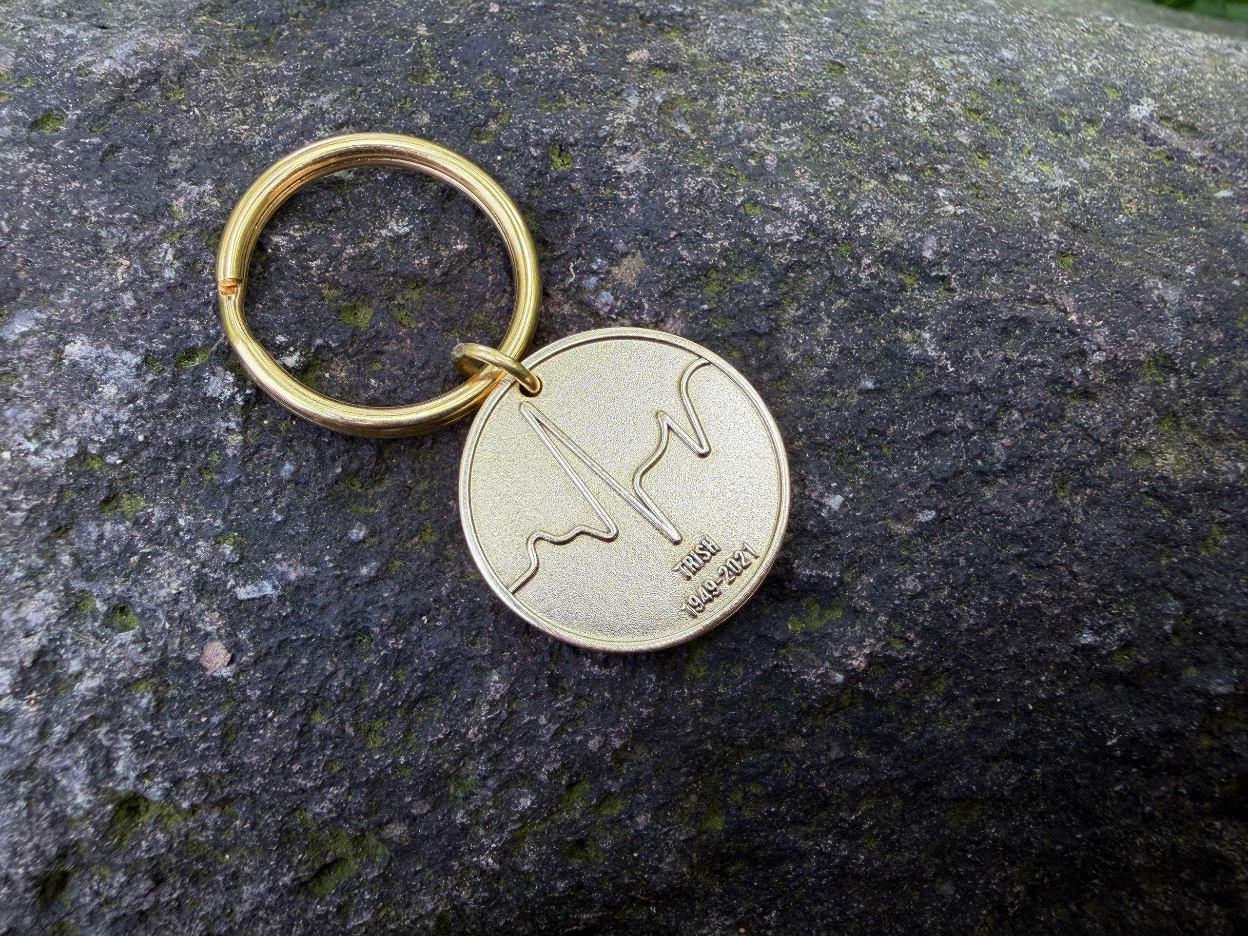 Gold keychain with wave engraving and text IRISH 1949-2021 on a stone surface.