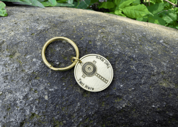 Gold keychain with Monstrance Cross design on a stone surface, surrounded by green leaves.