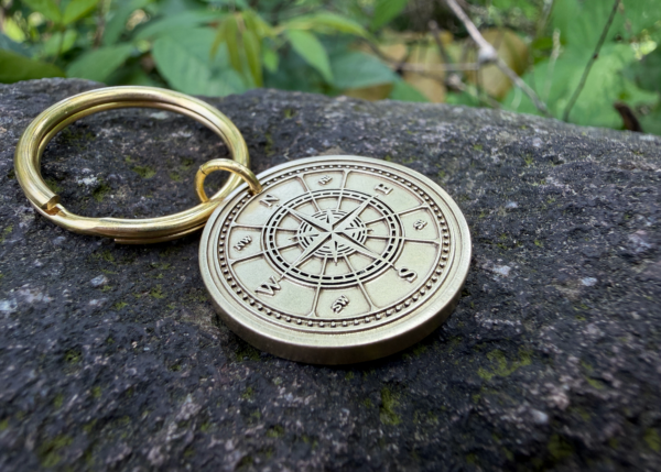 Gold keychain with engraved compass design on a rock surface, surrounded by green leaves.