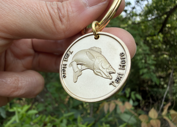 Gold keychain with engraved fish and customizable Text Here displayed against a leafy background.