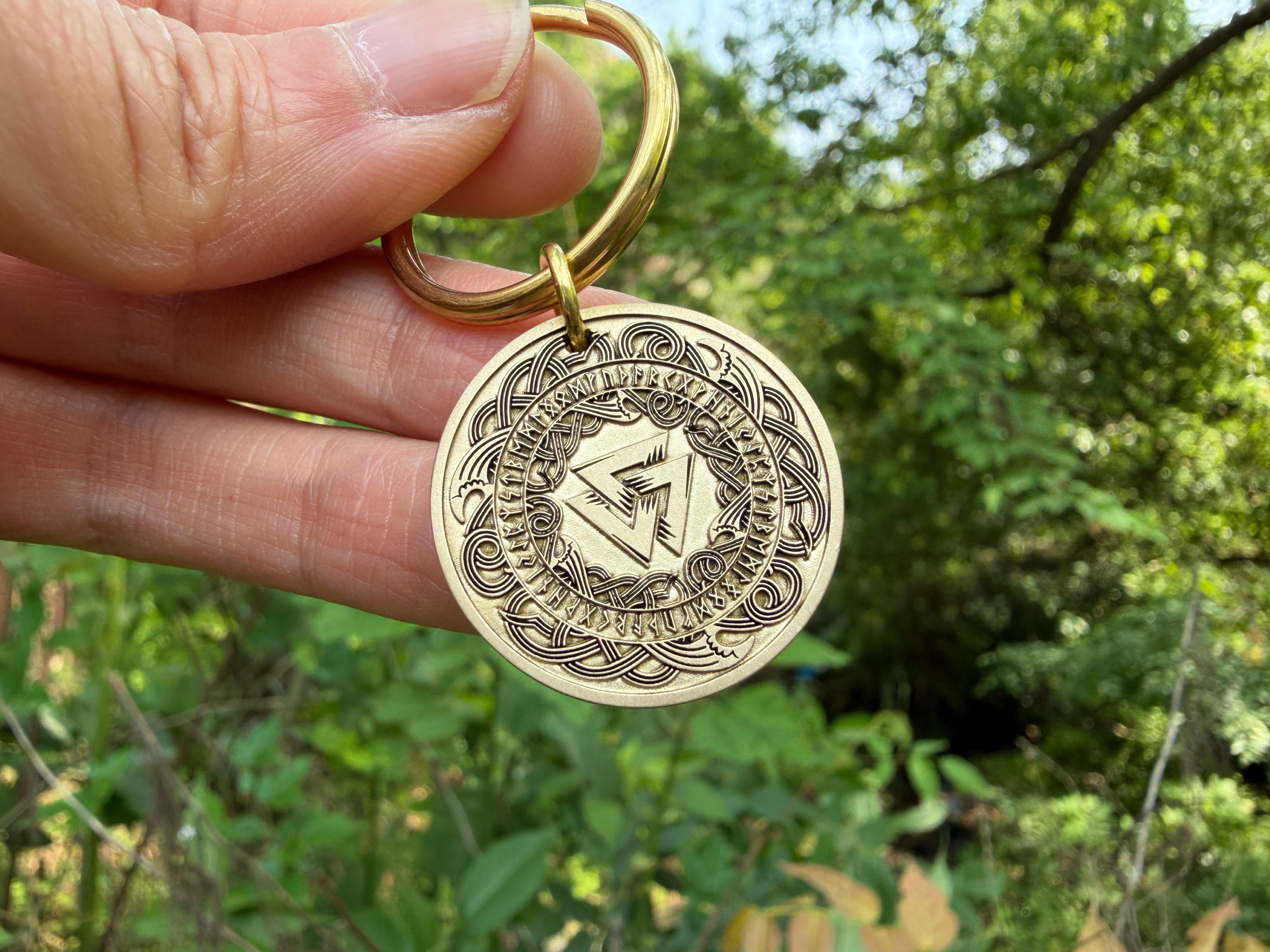 Person holding a gold keychain with intricate Norse runes and decorative Celtic patterns, set against a lush green background.