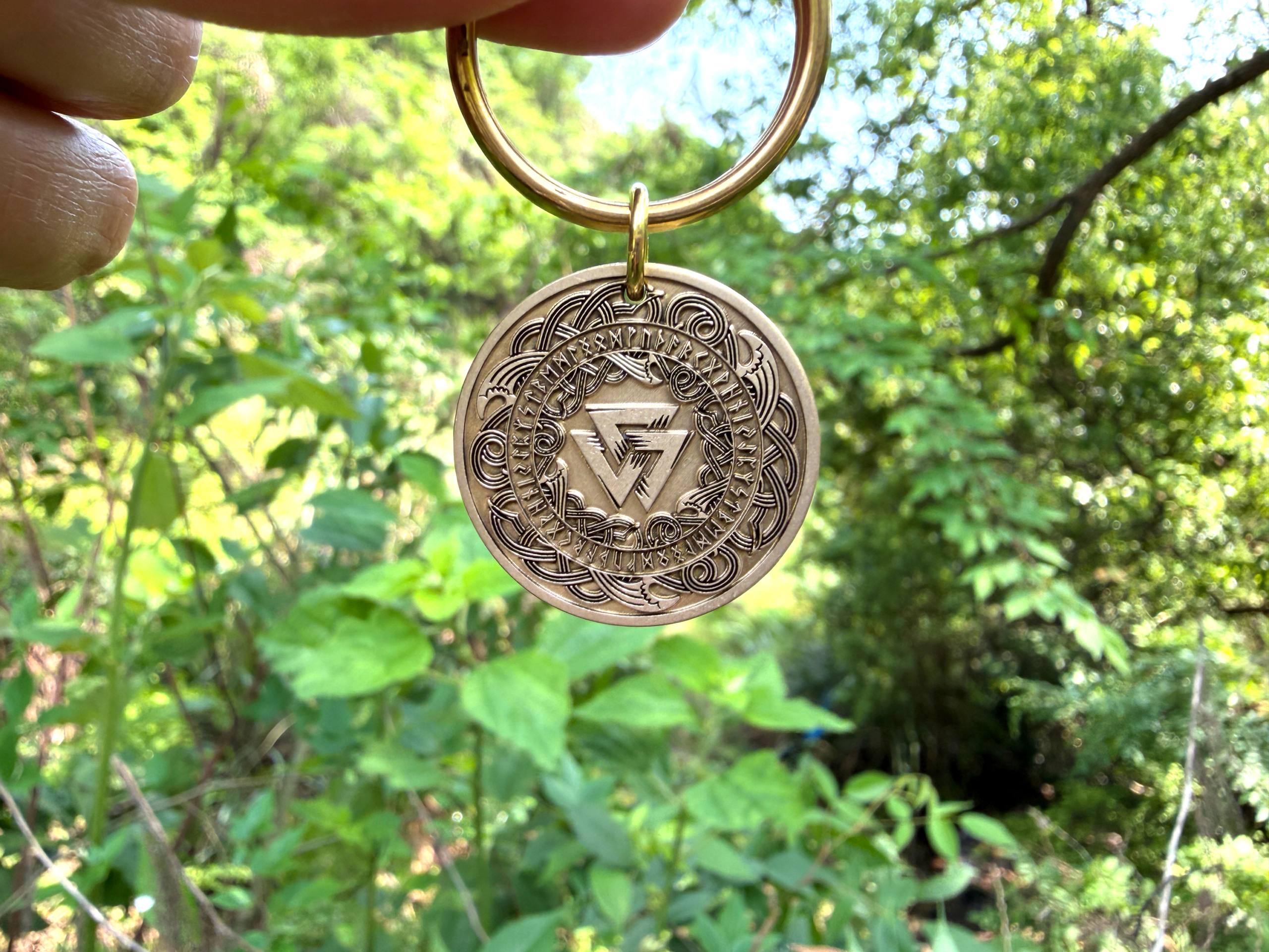 A golden keychain with a Vegvisir engraving held against a lush green forest background.