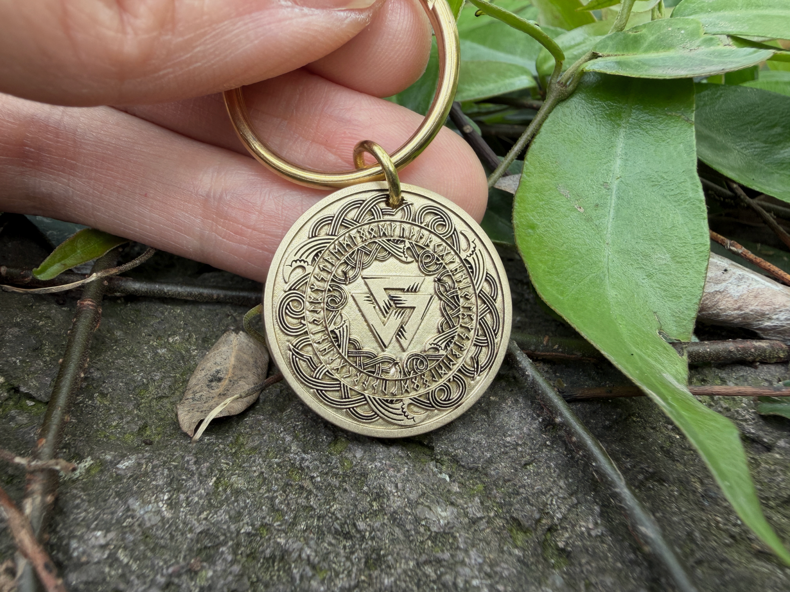 Gold keychain featuring intricate Norse runes and Viking symbols, held by hand over a natural background with leaves.
