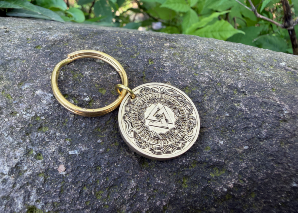 Golden keychain with intricate Norse design engraved on a coin, resting on a mossy rock in natural surroundings.