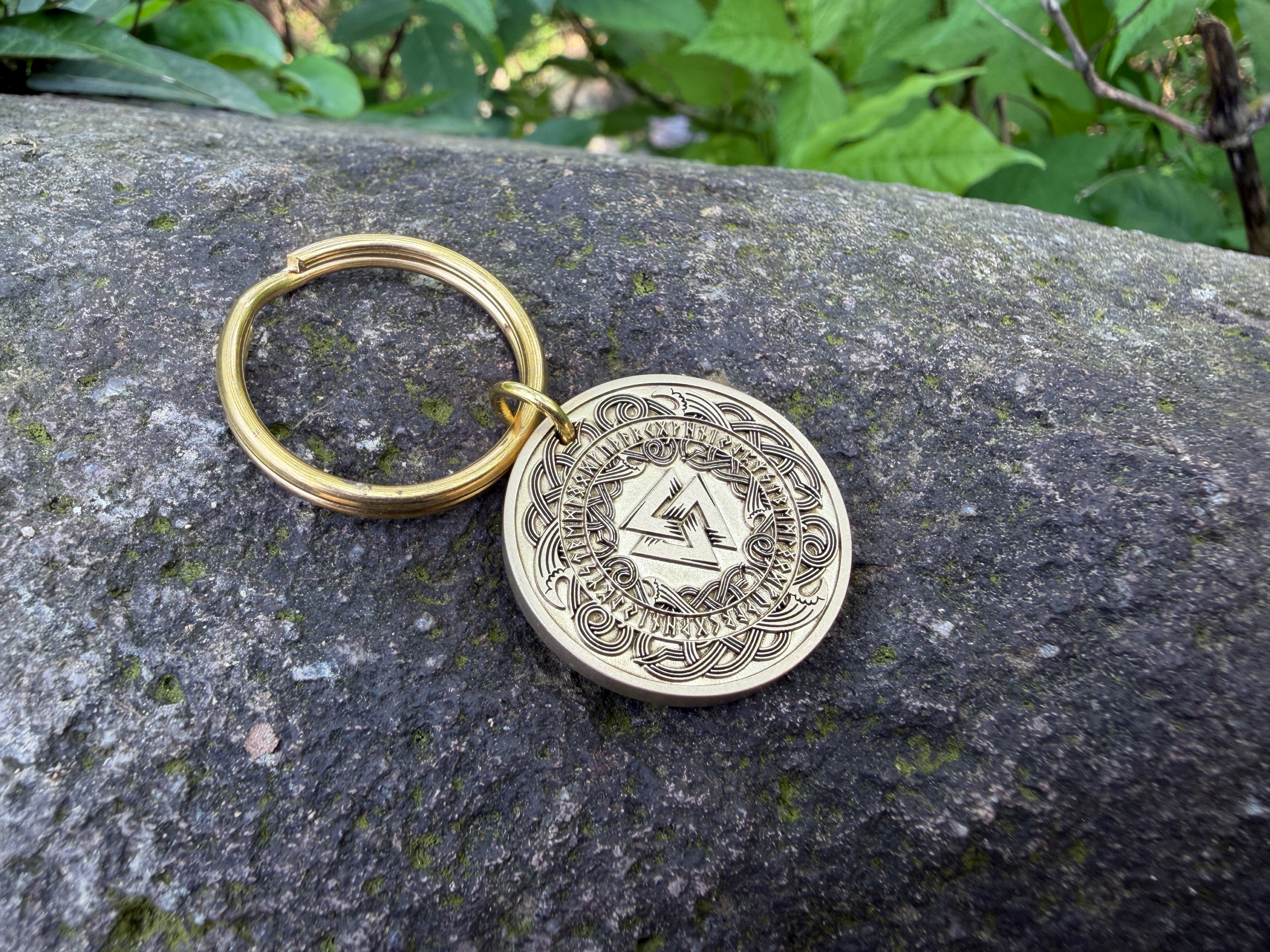 Golden keychain with intricate Norse design engraved on a coin, resting on a mossy rock in natural surroundings.