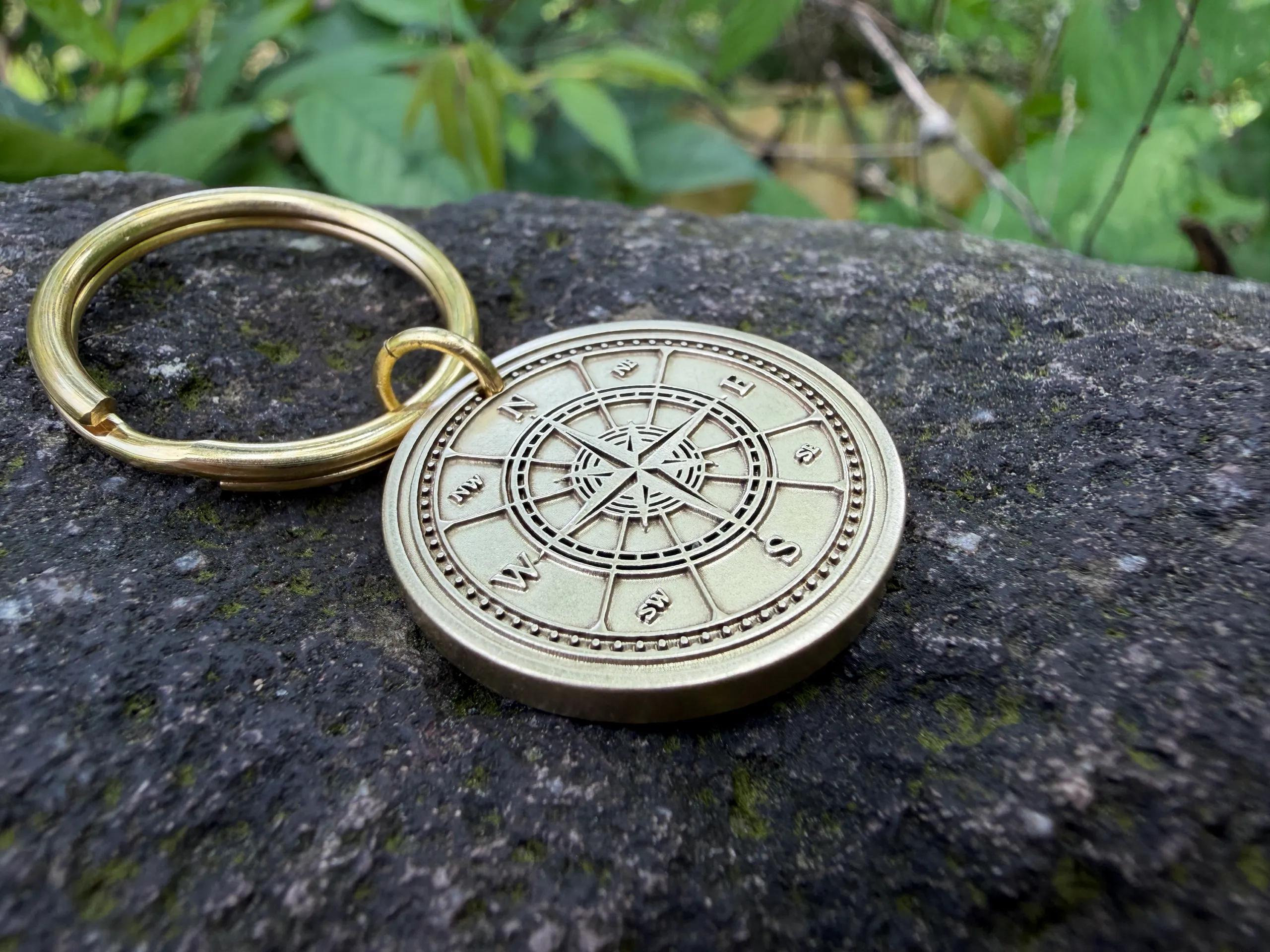 Gold keyring with engraved compass rose on stone background, surrounded by green leaves.