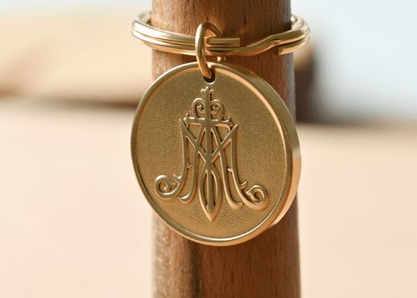 Gold circular keychain tag embossed with a Marian monogram featuring an M and a cross