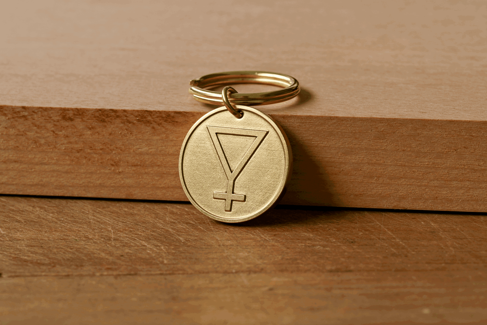 A circular brass keychain tag engraved with an inverted triangle with a cross below, resting on a wooden surface