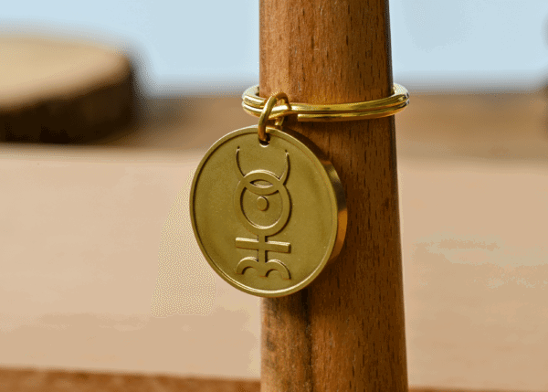Gold brass keychain tag engraved with an alchemical symbol, displayed on a wooden post