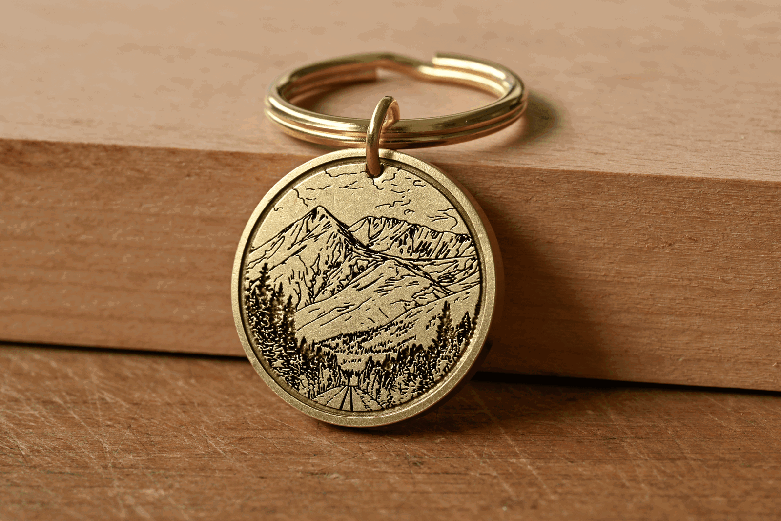 Brass keychain medallion with an engraved mountain landscape featuring pine trees and a road