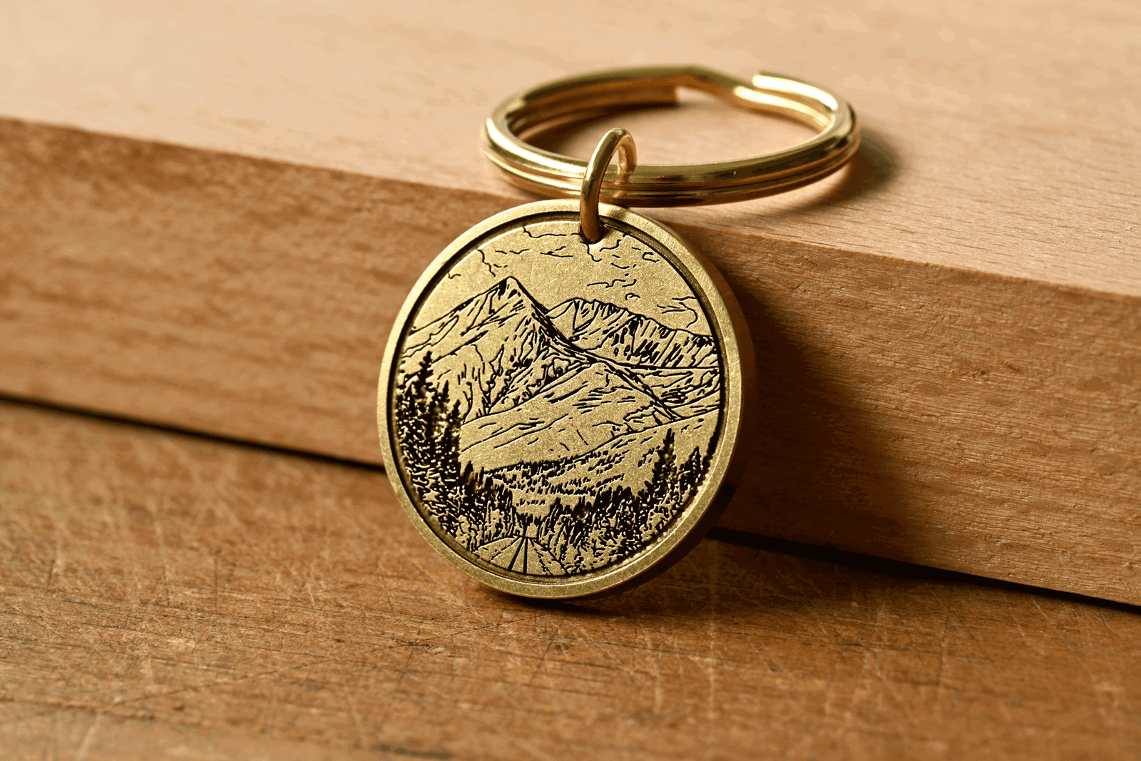 Round brass keychain engraved with a detailed mountain landscape and pine trees