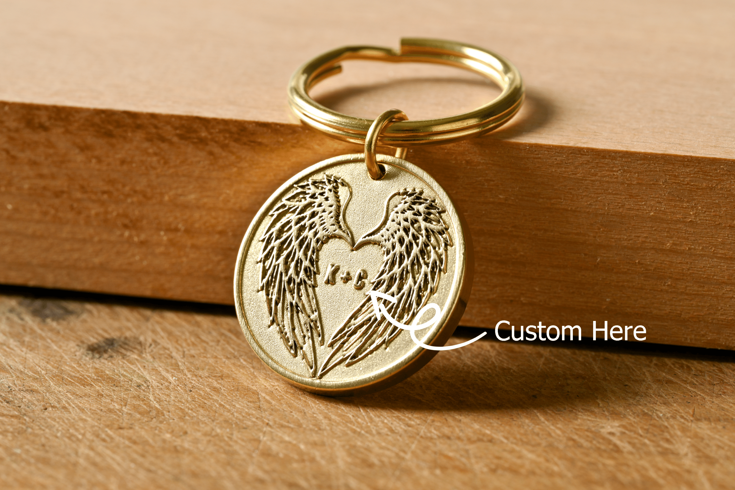 Gold round keychain engraved with angel wings forming a heart, with an arrow pointing to center initials labeled Custom Here