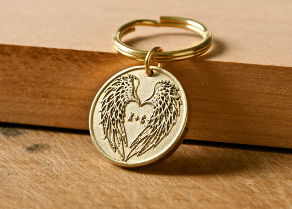 Round gold keychain engraved with angel wings forming a heart shape around the initials K + C