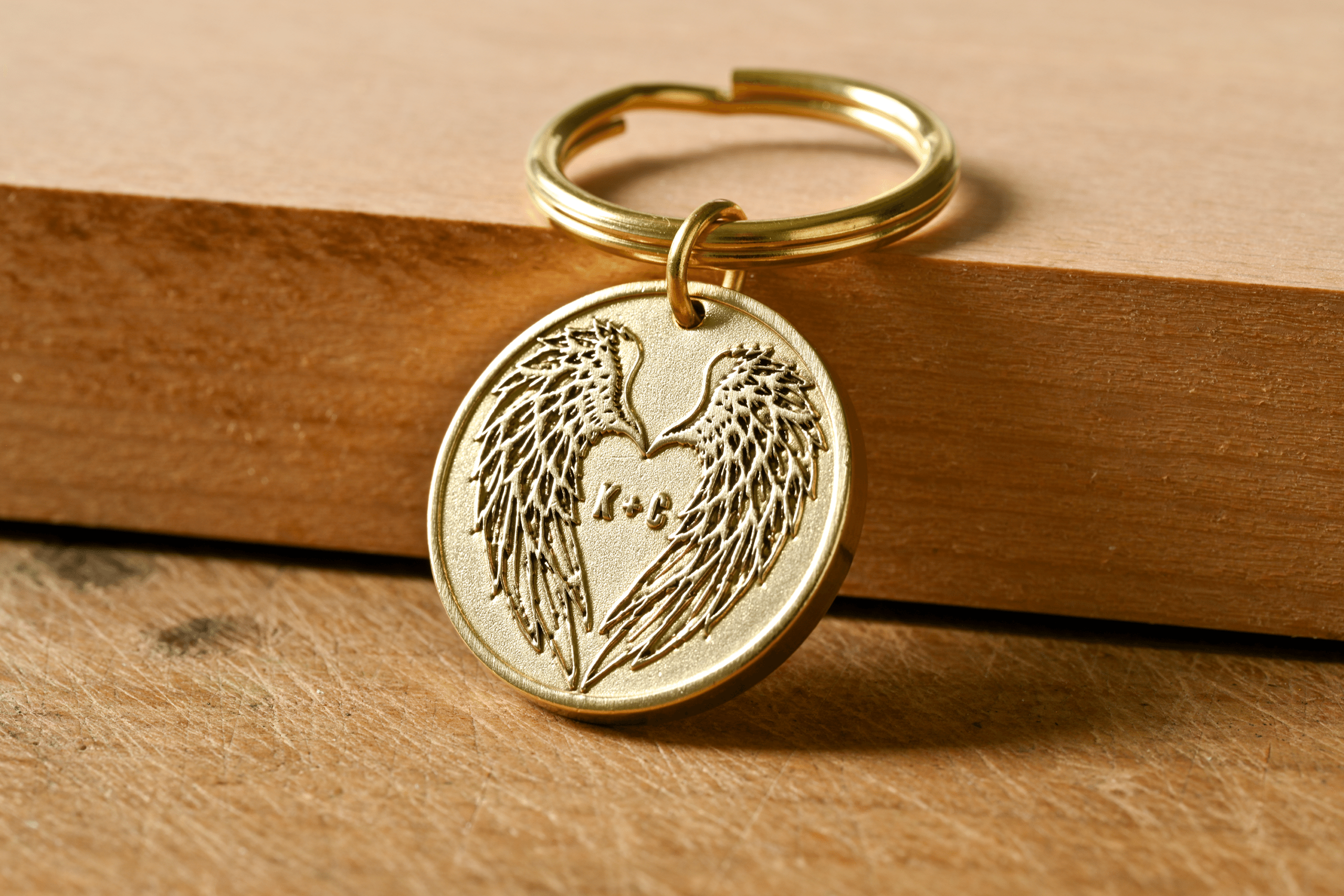 Round gold keychain engraved with angel wings forming a heart shape around the initials K + C