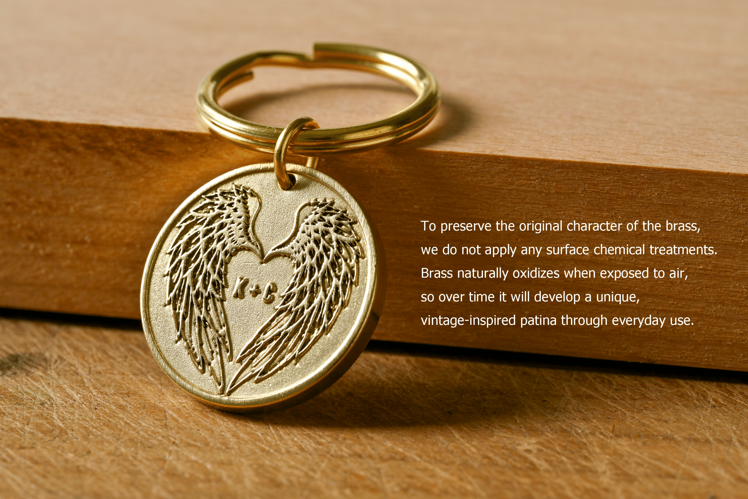 Brass keychain medallion engraved with angel wings forming a heart around "K+C". Text: To preserve the original character of the brass, we do not apply any surface chemical treatments. Brass naturally oxidizes when exposed to air, so over time it will develop a unique, vintage-inspired patina through everyday use
