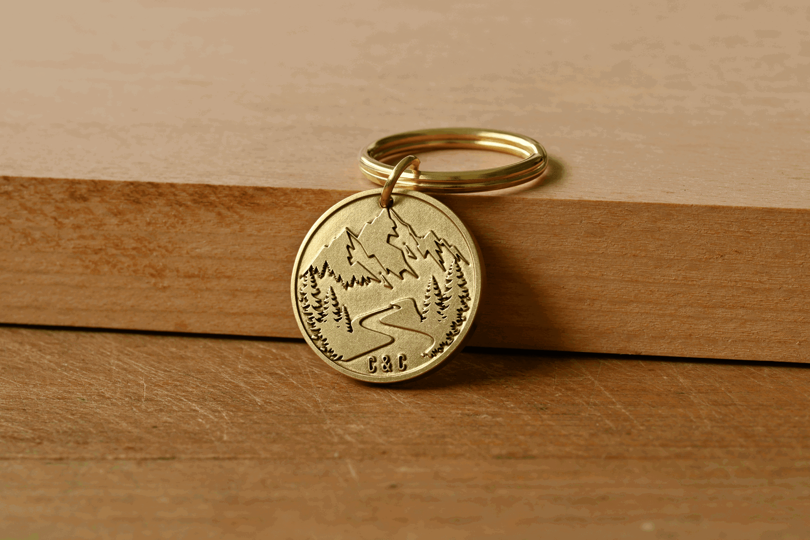 Brass keychain featuring an engraved mountain landscape with trees, a winding path, and the initials C&C stamped at the bottom