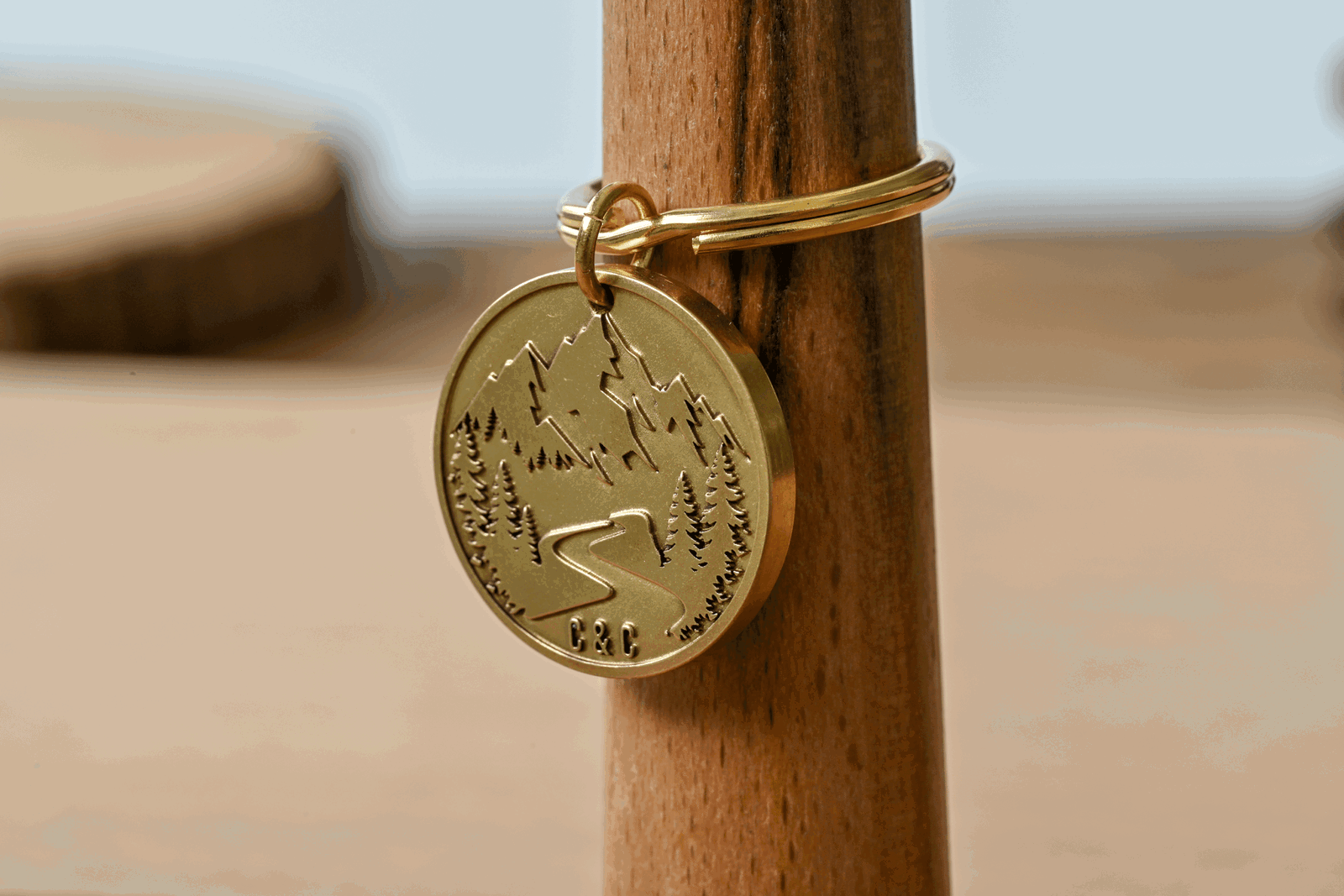 Gold circular keychain tag engraved with a mountain landscape, pine trees, winding road, and initials C & C