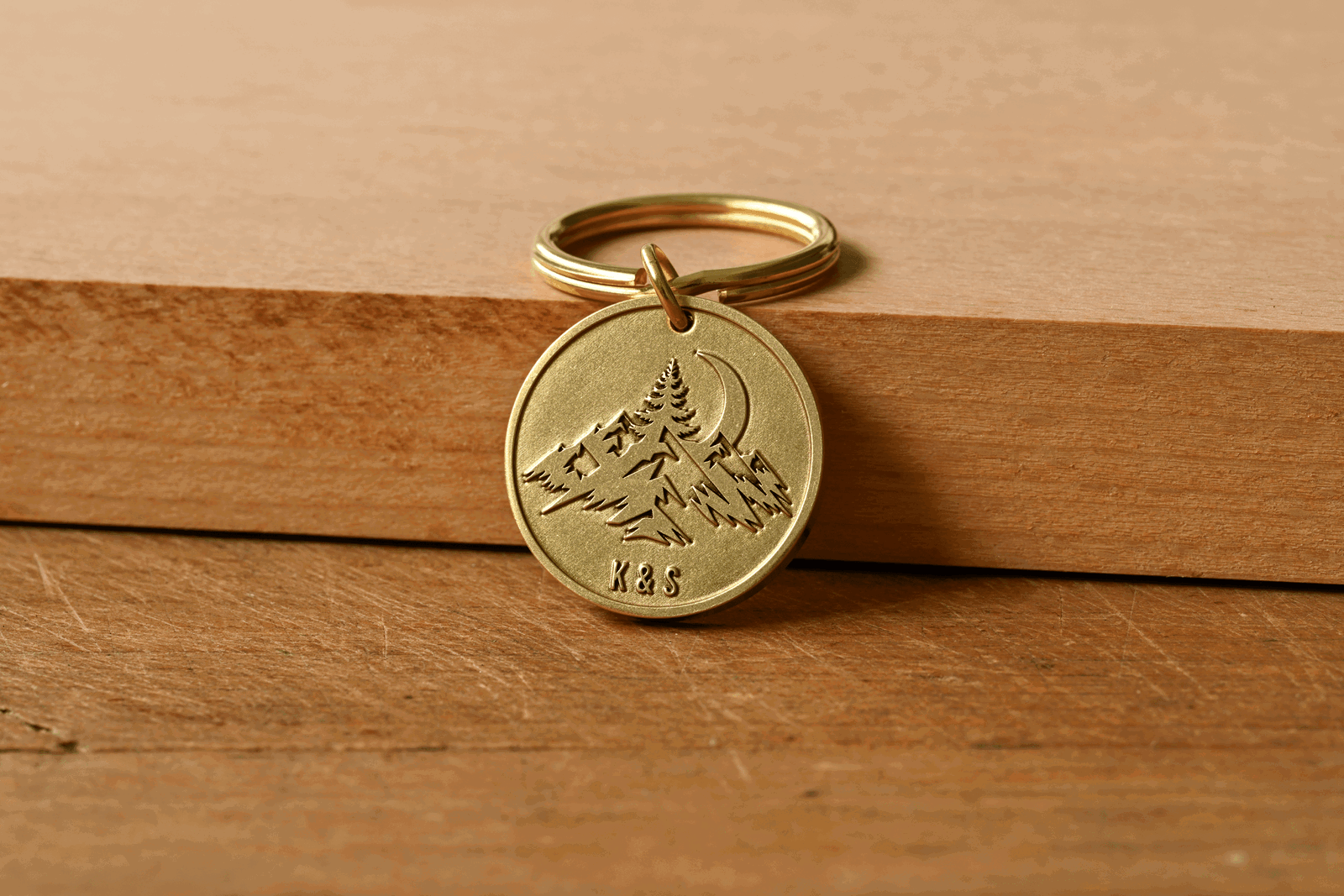 Round brass keychain engraved with mountain peaks, pine trees, a crescent moon, and the initials K&S